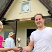 Janet Bazley presents The Colin Bazley Single Wicket Cup to the first winner, Dan Carter. Behind them is the rededicated Clavering Cricket Club pavilion, with the sign reading: The Colin Bazley Pavilion
